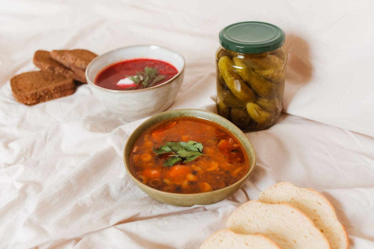 Illustrative stock photo: bowls of borscht and solyanka soup beside a jar of pickles
