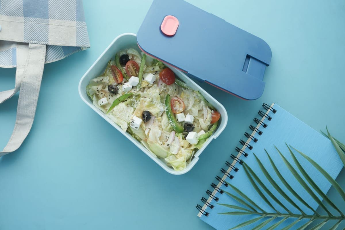 A salad lunch box beside a notebook on a blue background, seen from above