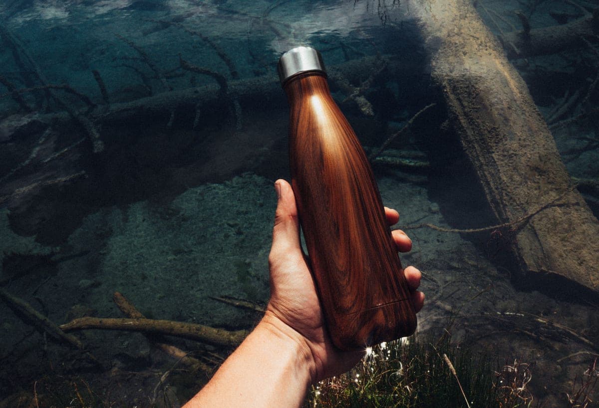 A hand holding a stainless steel water bottle over a stream